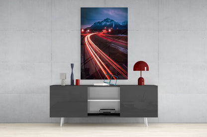 Road Glass Wall Art displayed above a gray media console, showing a long-exposure photograph of red car light trails curving toward a snow-capped mountain at dusk.