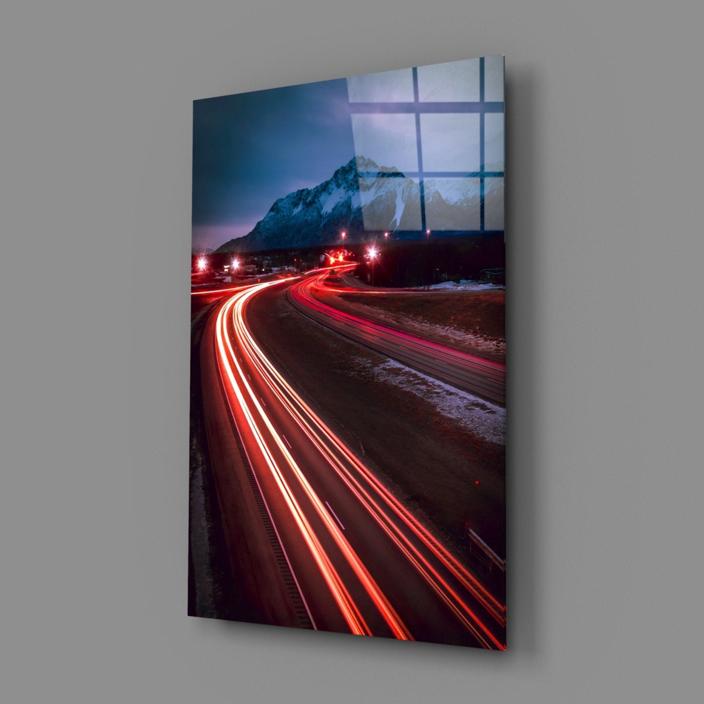 Road Glass Wall Art displaying long-exposure car light trails in red against a snowy mountain landscape at dusk, mounted on gray wall.