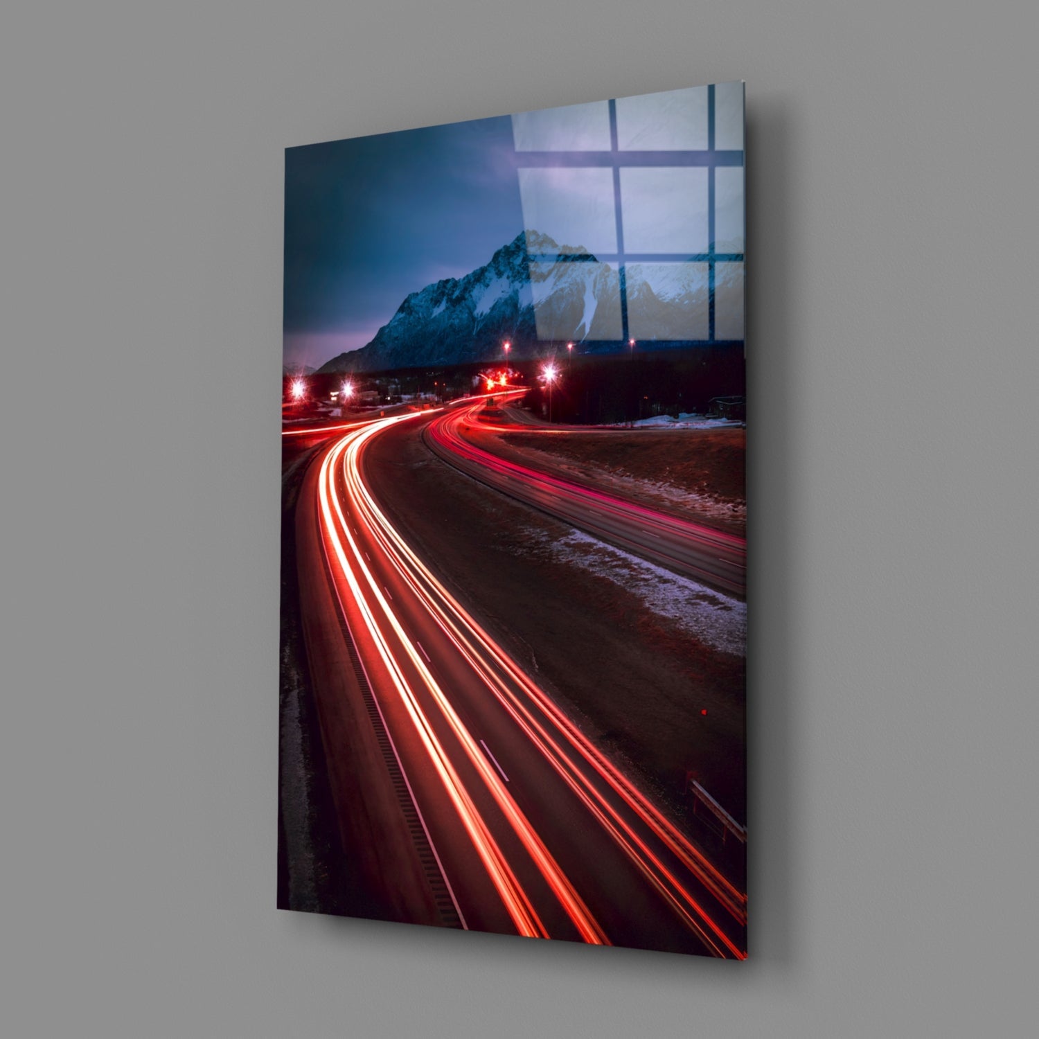 Road Glass Wall Art displaying long-exposure car light trails in red against a snowy mountain landscape at dusk, mounted on gray wall.