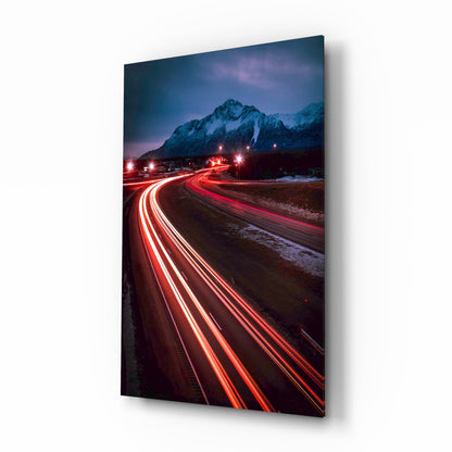 Angled product display of a glass wall art panel showing a nighttime mountain road with red car light trails curving toward snow-capped peaks under twilight sky.