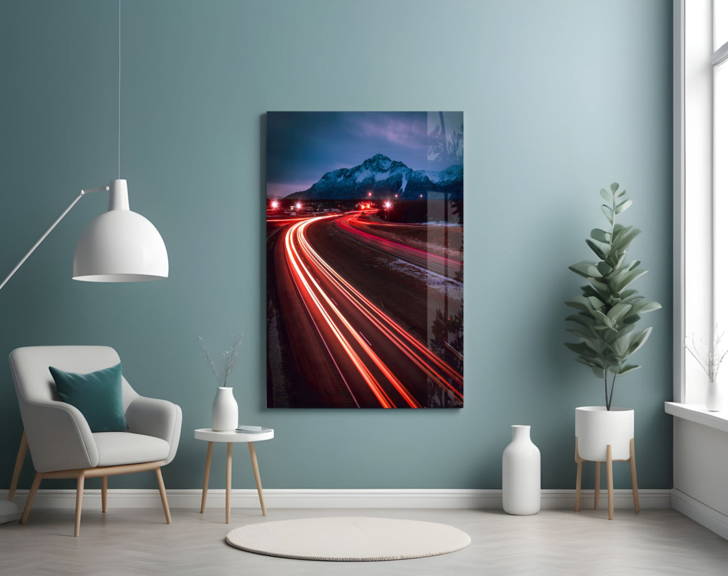 Road Glass Wall Art displayed on teal wall above white chair, showing long-exposure light trails on mountain road at dusk.