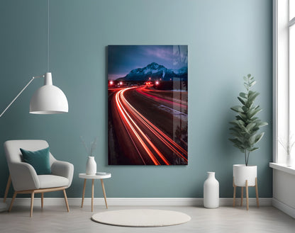Road Glass Wall Art displayed on teal wall above white chair, showing long-exposure light trails on mountain road at dusk.