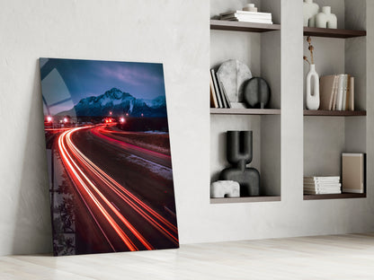 Glass wall art leaning against white wall displaying red car light trails and snowy mountain at twilight.