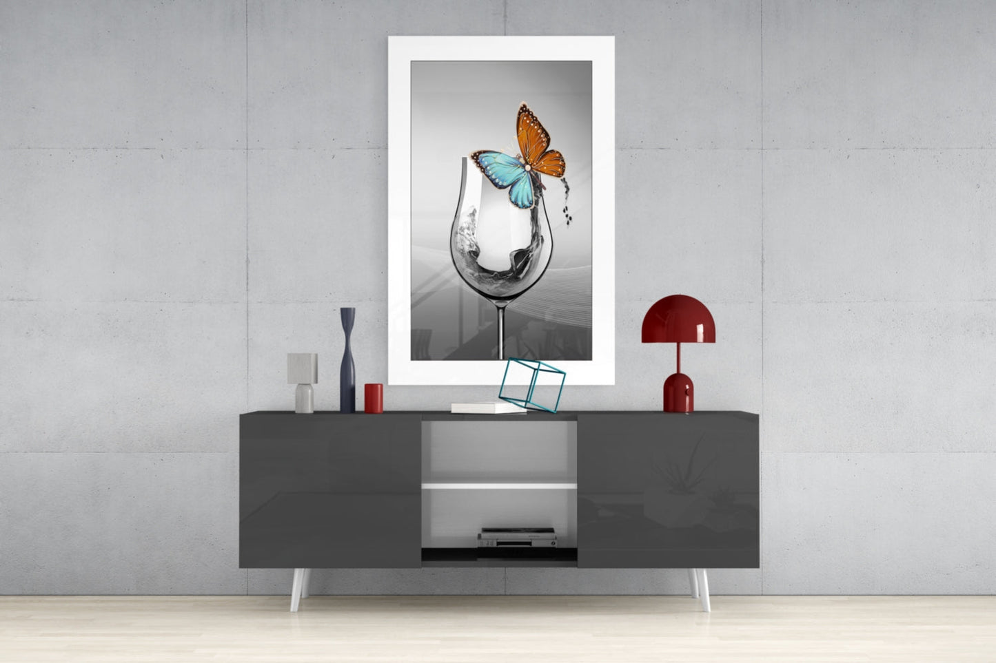 Framed wall art displaying a wine glass with water splash and orange-blue butterfly, hung above gray console table with red lamp and decorative objects.