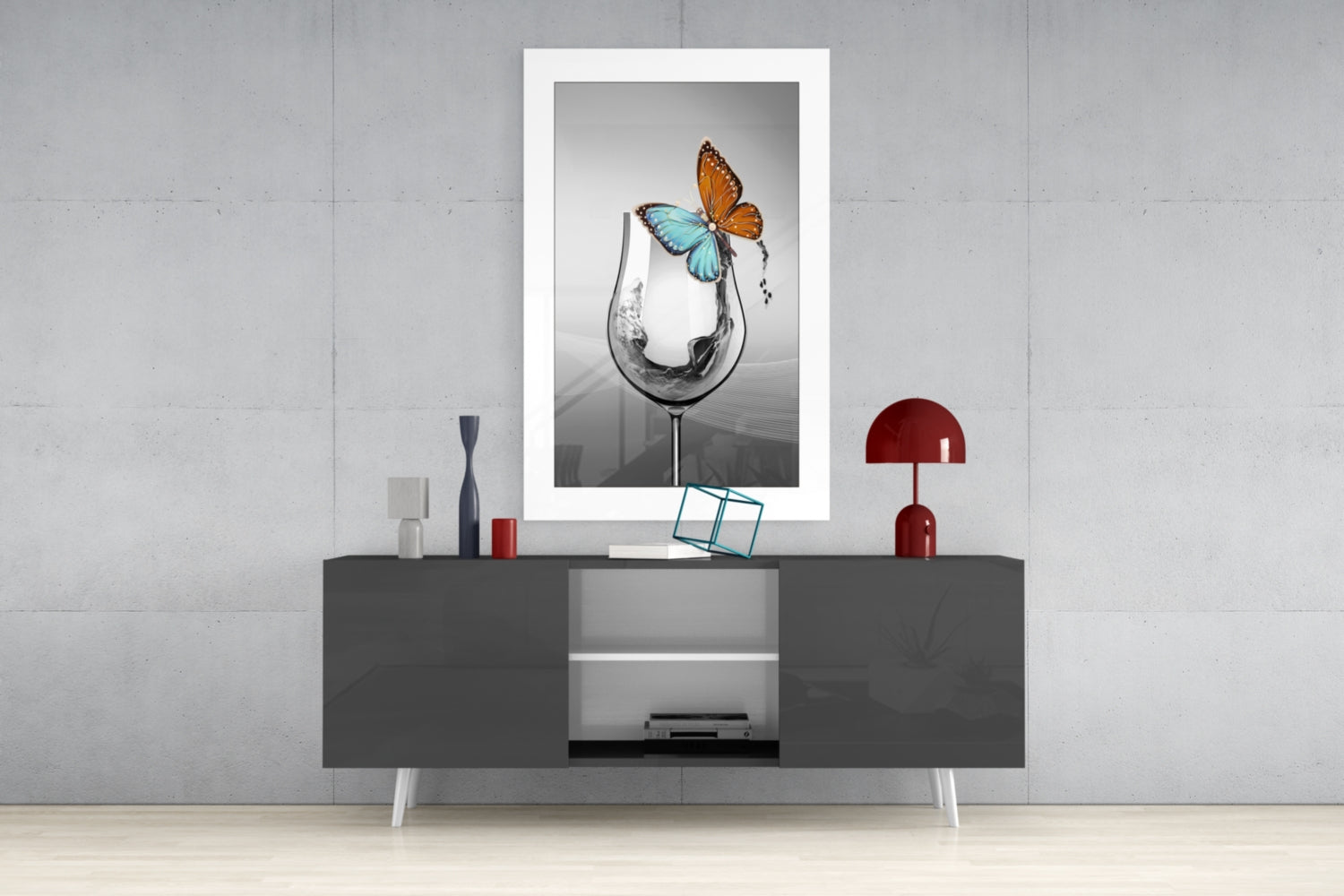 Framed wall art displaying a wine glass with water splash and orange-blue butterfly, hung above gray console table with red lamp and decorative objects.