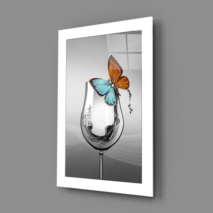 Framed white-bordered wall art displaying a black and white wine glass with colorful blue and orange butterflies perched on the rim, angled perspective view.