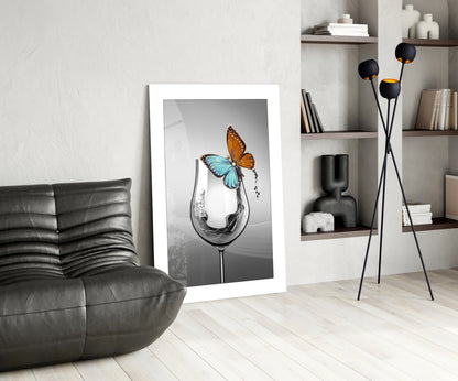 Framed wall art depicting a wine glass with water splash and colorful butterflies, displayed in modern living room setting with leather sofa and shelving.
