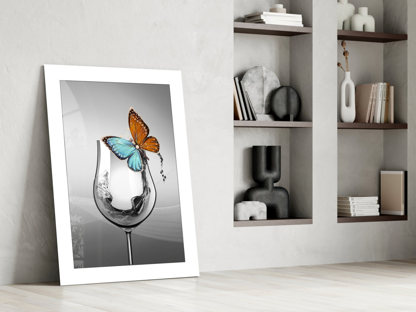 Framed white-bordered art print leaning against wall depicting a wine glass with splashing water and colorful butterfly, displayed in minimalist interior setting.