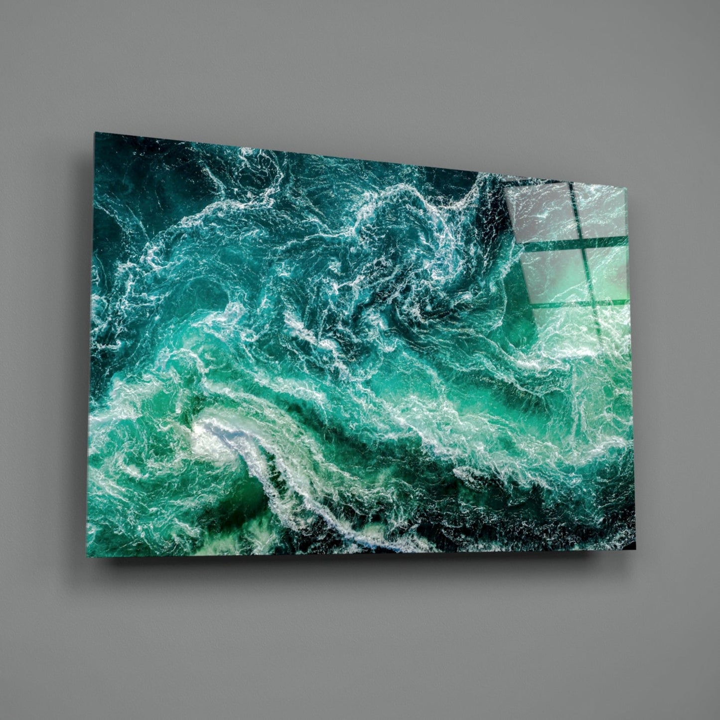 Angled view of glass wall art displaying turbulent teal and white ocean waves with a dark rocky coastline visible.