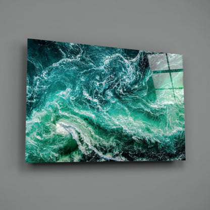 Angled view of glass wall art displaying turbulent teal and white ocean waves with a dark rocky coastline visible.