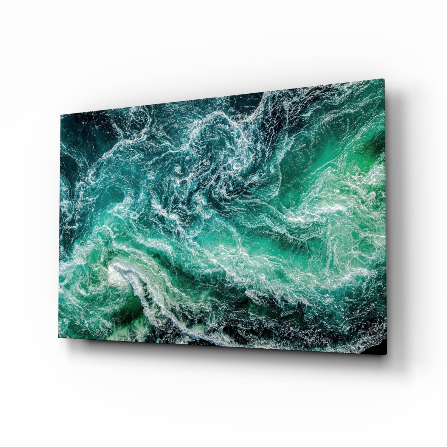 Angled view of glass wall art displaying aerial ocean waves in shades of green and white with foam patterns.