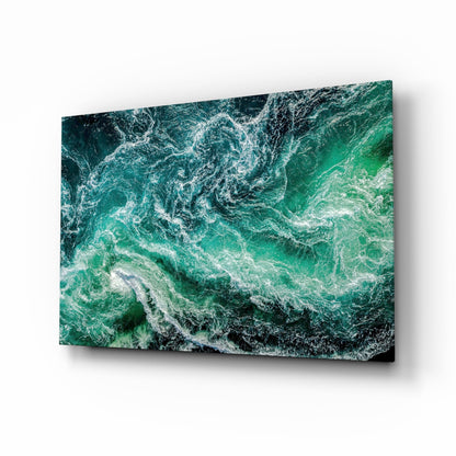 Angled view of glass wall art displaying aerial ocean waves in shades of green and white with foam patterns.