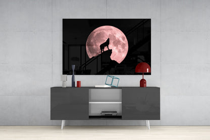 Glass wall art depicting a silhouetted wolf howling on a rocky outcrop against a large pink full moon, displayed on a gray media console.