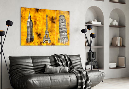 Three-panel glass wall art displaying Big Ben, Eiffel Tower, and Leaning Tower of Pisa on vibrant yellow textured background, hung above black leather sofa in modern living room.