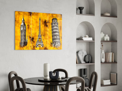 Three-panel glass wall art displaying Big Ben, Eiffel Tower, and Leaning Tower of Pisa on vibrant yellow background, hung above dining table.
