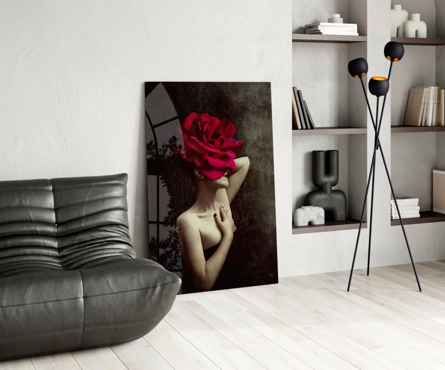 Modern living room with gray leather chair, glass wall art depicting woman's face covered by large red rose, black tripod floor lamp, and white shelving unit.