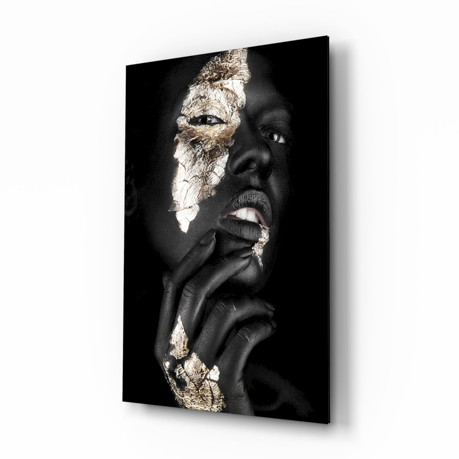Portrait of a person with dark skin and gold leaf accents on face and hands against black background, displayed as glass wall art.
