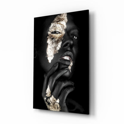 Portrait of a person with dark skin and gold leaf accents on face and hands against black background, displayed as glass wall art.