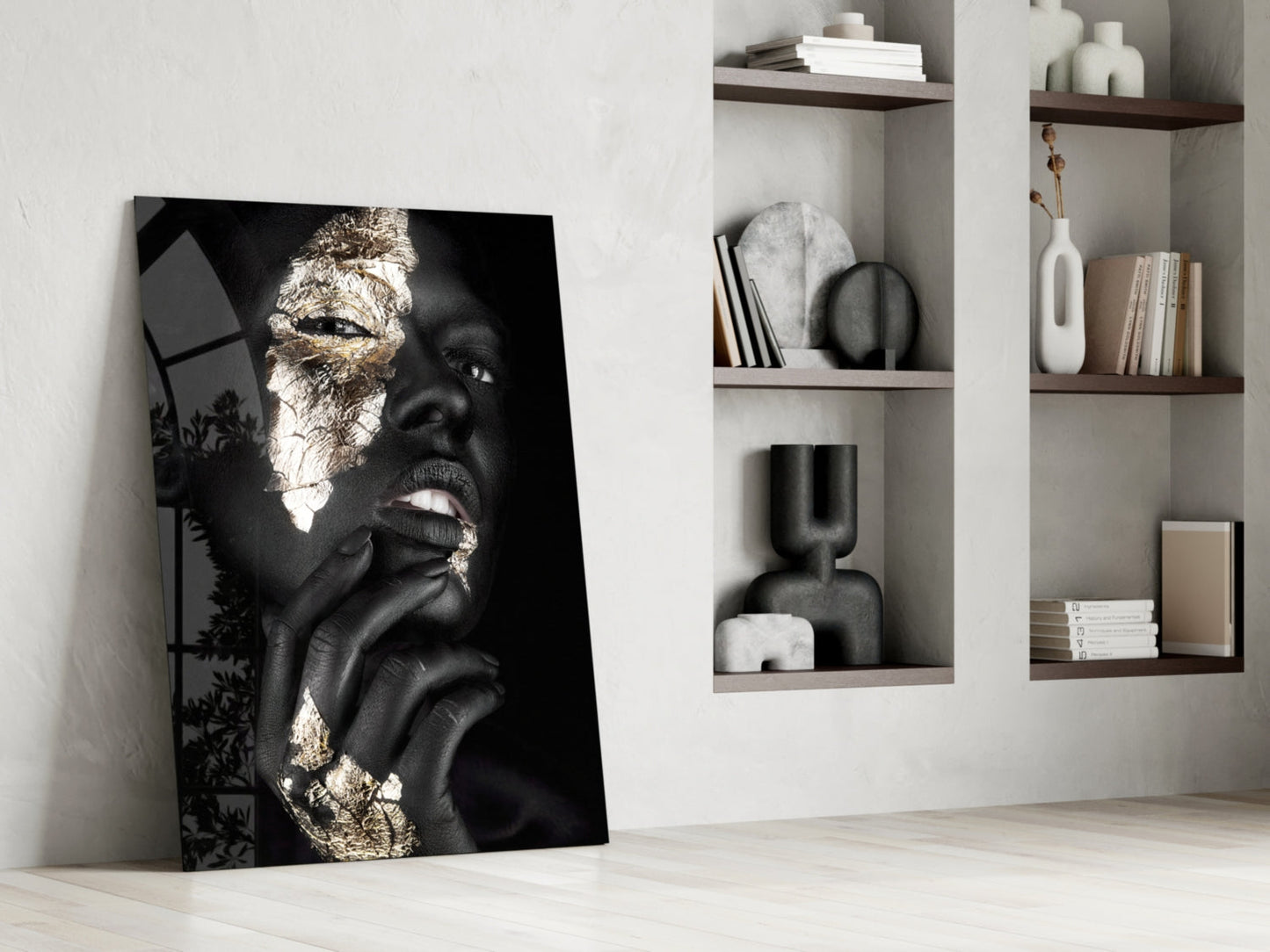 Black and gold portrait glass art leaning against white wall beside styled shelving unit with decorative objects and books.