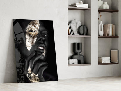 Black and gold portrait glass art leaning against white wall beside styled shelving unit with decorative objects and books.