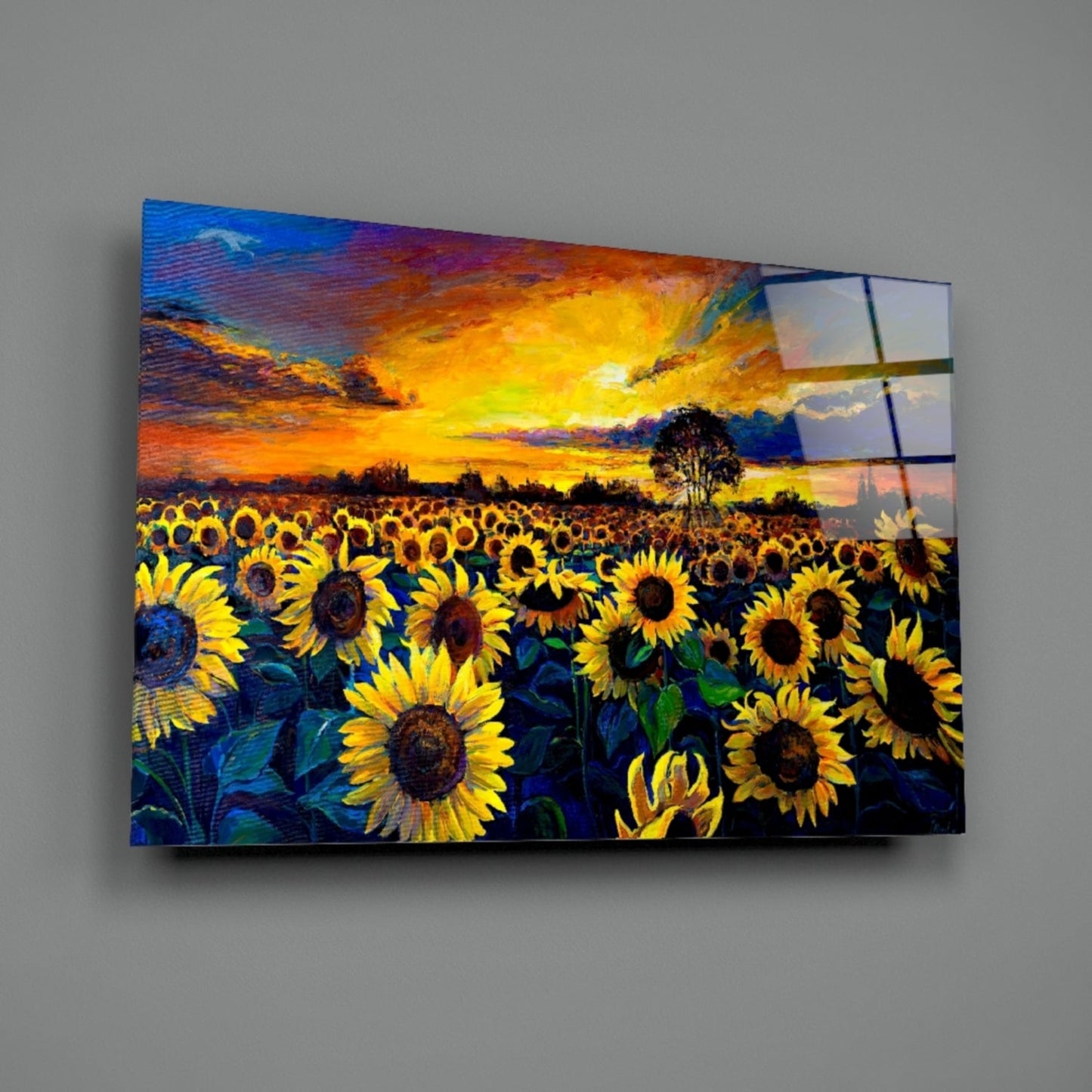 Glass wall art panel mounted on gray wall displaying vibrant sunflower field at sunset with blue sky and silhouetted tree.