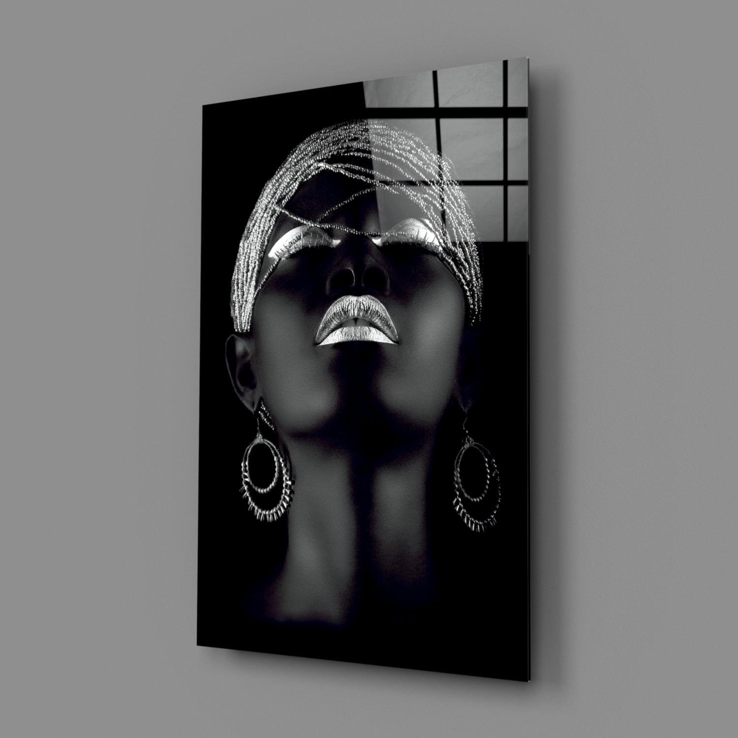 Angled view of black glass wall art featuring silver-highlighted portrait of woman wearing sunglasses, wrap headwrap, and ornamental earrings against grey background.