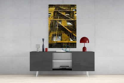 Yellow metal fire escape stairs on building facade displayed as vertical glass wall art above gray media console.