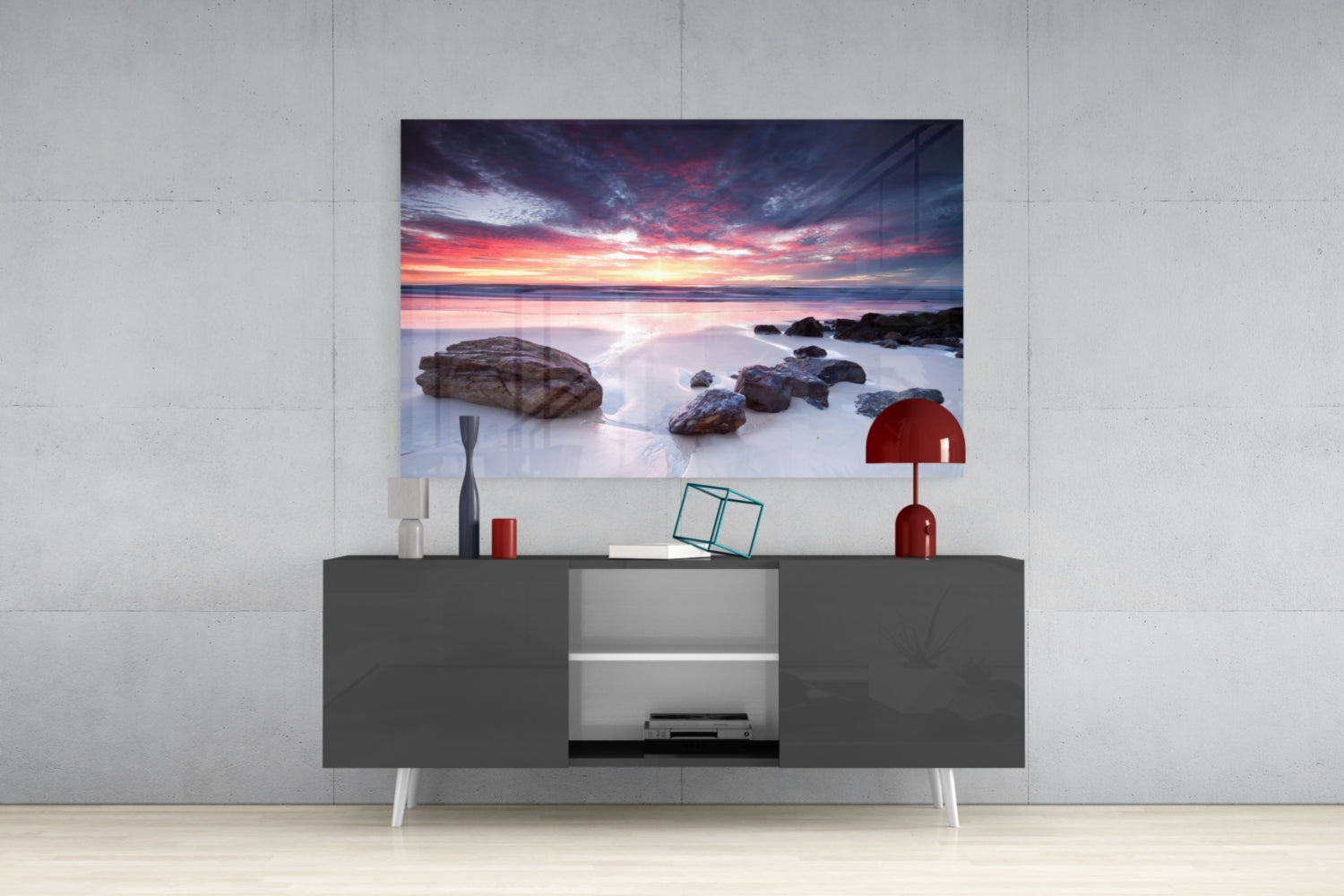 Glass wall art depicting a dramatic sunset over rocky beach with calm water, displayed above dark gray media console in modern interior.