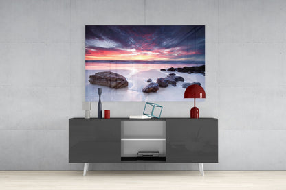 Glass wall art depicting a dramatic sunset over rocky beach with calm water, displayed above dark gray media console in modern interior.