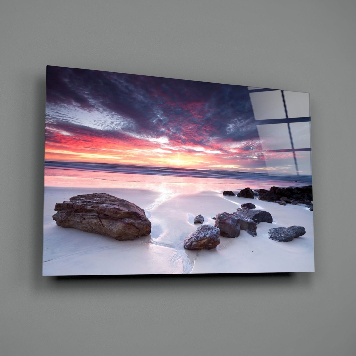 Mounted glass wall art showing a beach sunset with rocks, pink and orange sky reflected on a gray wall, displaying dark grid lines across the surface.