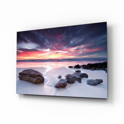 Mounted glass wall art displaying a dramatic coastal sunset with vibrant pink and orange sky over rocky beach and calm ocean waters.