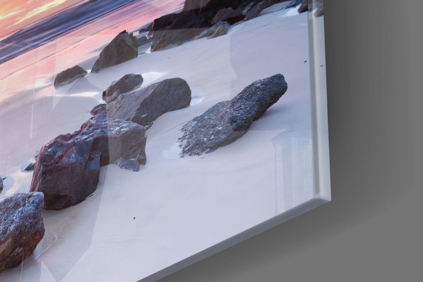 Angled view of glass wall art displaying snow-covered rocks and beach scene with pink sunset sky reflection on white frame.