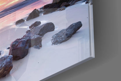 Angled view of glass wall art displaying snow-covered rocks and beach scene with pink sunset sky reflection on white frame.