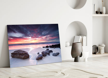 Sunset beach scene with rocky shoreline displayed as framed glass art leaning against white wall in minimalist interior.