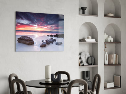 Sunset beach scene with golden sky and rocks on glass wall art displayed above round black dining table in modern interior.