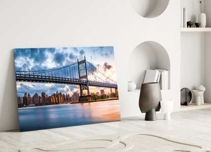 Glass wall art leaning against white wall displays illuminated bridge and city skyline at sunset in blue and orange tones.