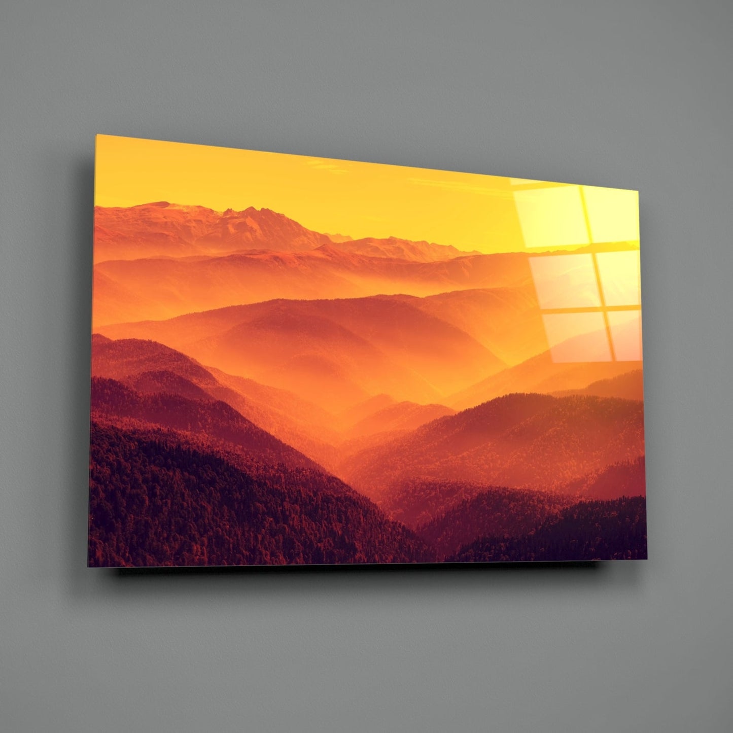 Angled view of glass wall art displaying layered mountains at sunset with warm orange and yellow tones, mounted on gray wall.