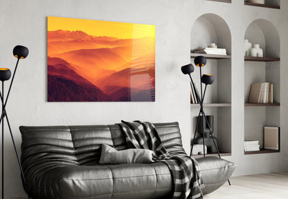 Warm Landscape Glass Wall Art mounted above black leather sofa, displaying golden sunset over layered mountains in living room setting.