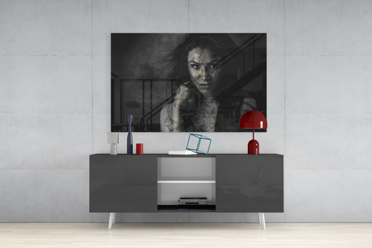 Black and white glass wall art of tattooed woman displayed above gray media console with red lamp and accessories in modern interior setting.