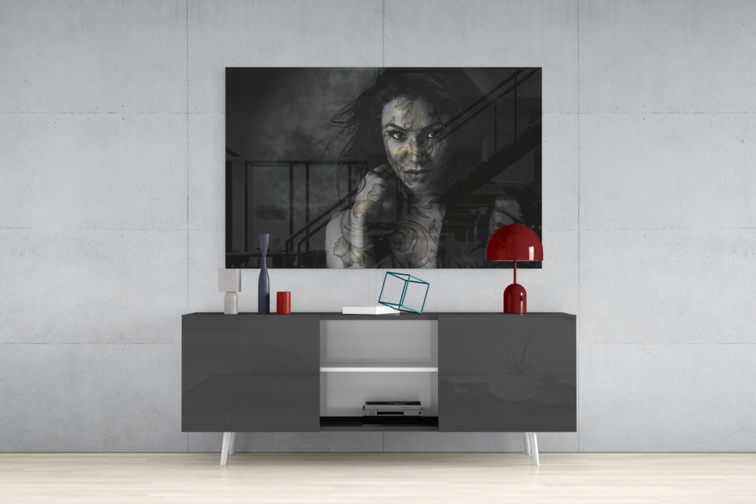 Black and white glass wall art of tattooed woman displayed above gray media console with red lamp and accessories in modern interior setting.