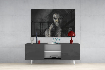 Black and white glass wall art of tattooed woman displayed above gray media console with red lamp and accessories in modern interior setting.