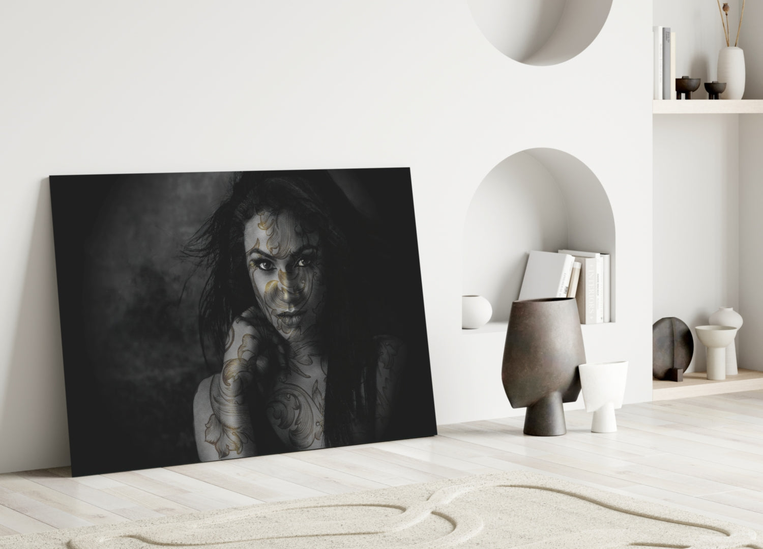 Black and white portrait of tattooed woman leaning against white wall in minimalist interior with shelving and decorative objects.