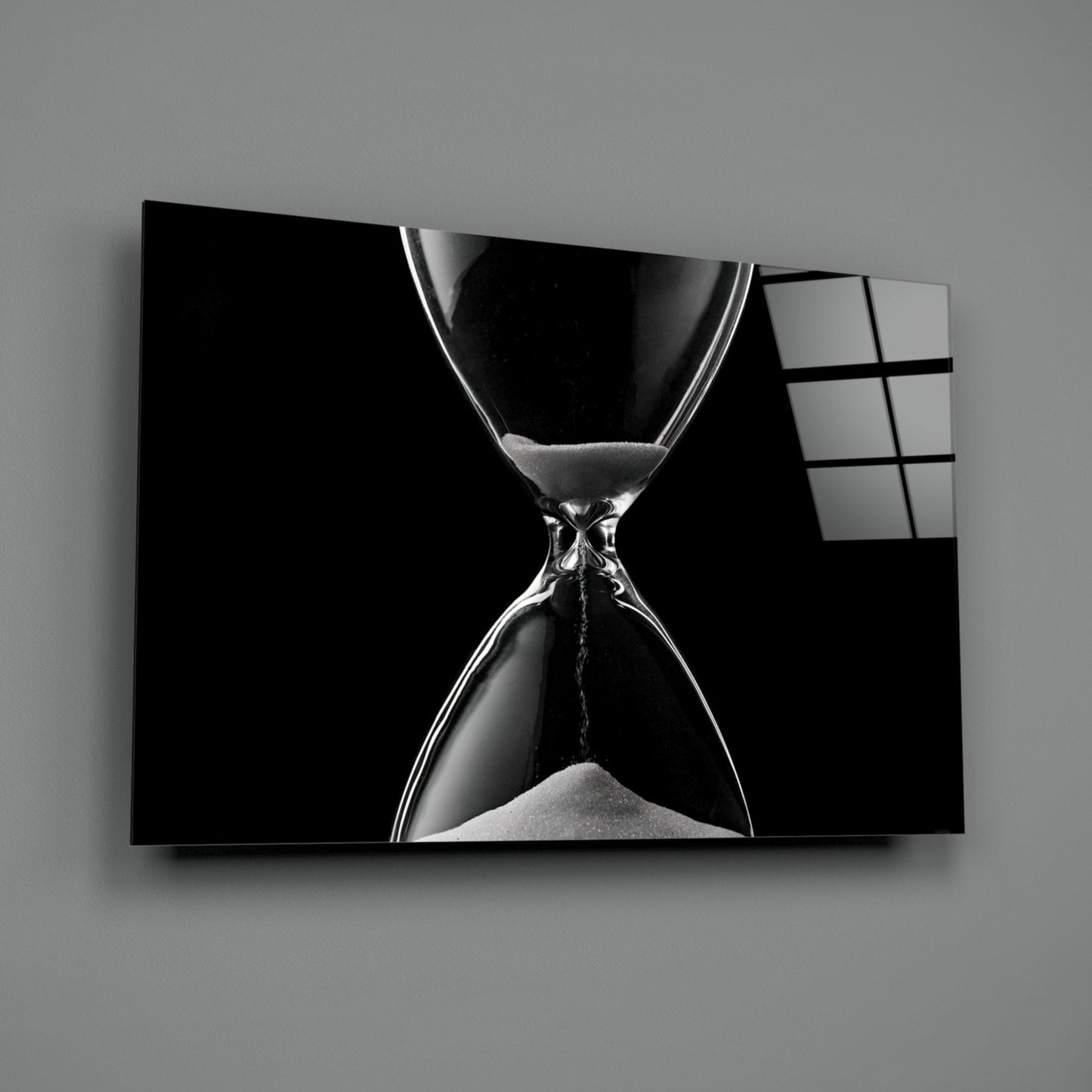 Black and white hourglass with flowing sand displayed on a wall-mounted glass panel against gray background.