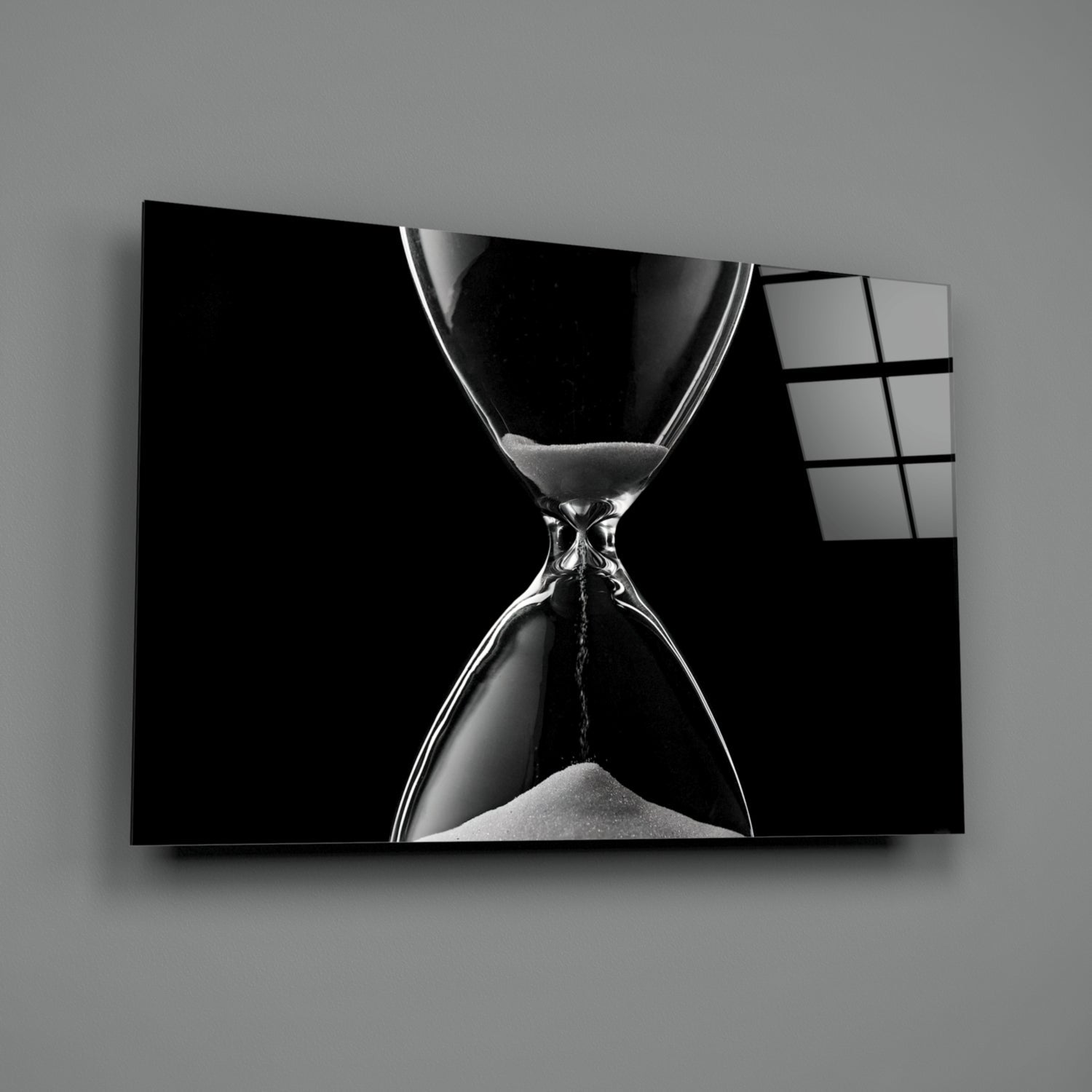 Black and white hourglass with flowing sand displayed on a wall-mounted glass panel against gray background.