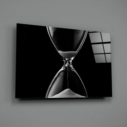 Black and white hourglass with flowing sand displayed on a wall-mounted glass panel against gray background.