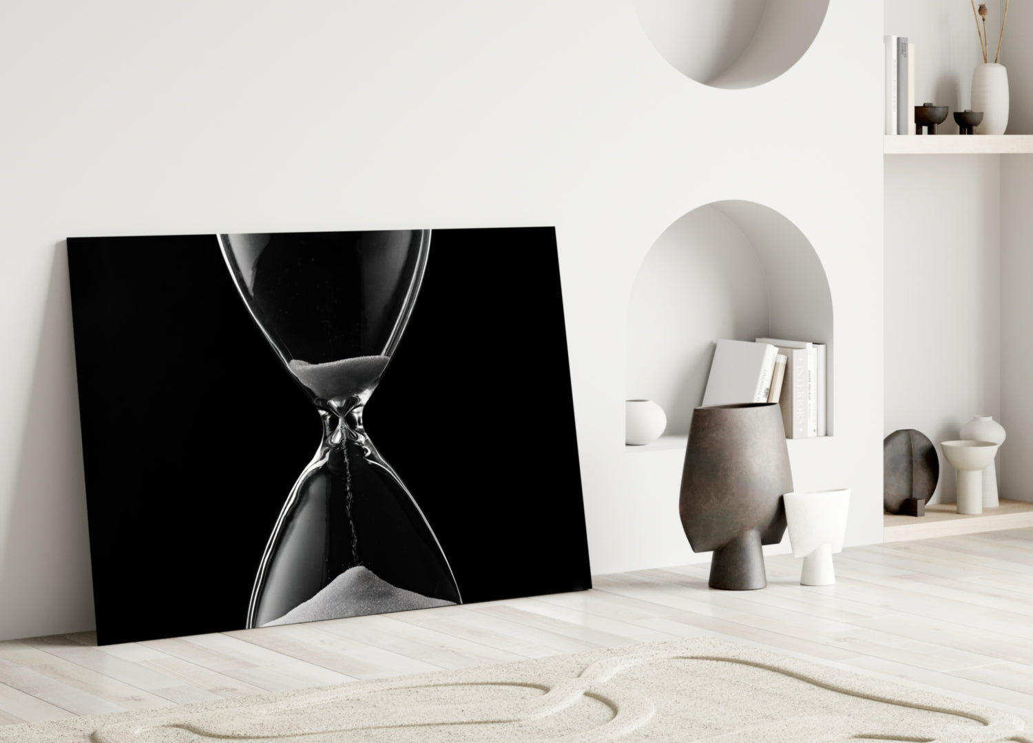 Black and white hourglass canvas leaning against white wall in minimalist room setting with sand and decorative objects.