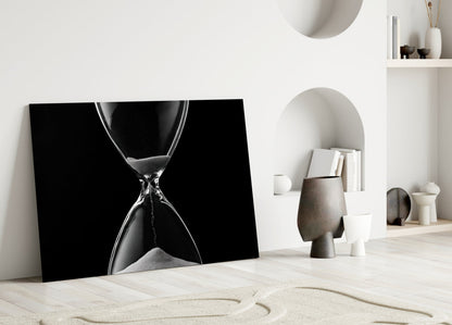 Black and white hourglass canvas leaning against white wall in minimalist room setting with sand and decorative objects.