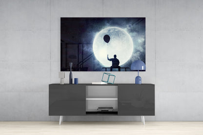 Glass wall art depicting silhouetted figure holding balloon against large glowing moon, displayed above dark media console in modern interior.