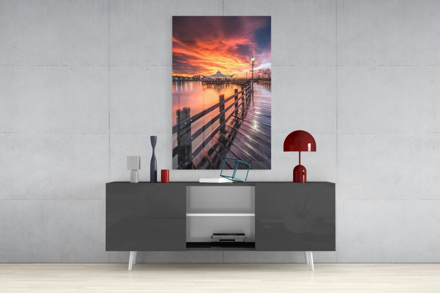 Bridge of Love Glass Wall Art displaying a wooden pier at sunset with orange and pink sky, mounted above a gray media console with decorative items.