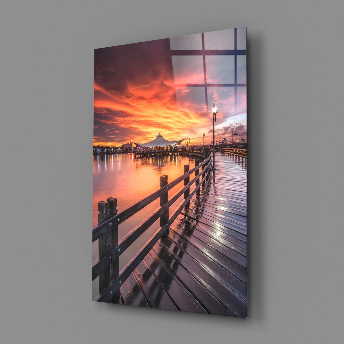 Glass wall art displaying a wooden pier extending toward a dramatic orange sunset over water, with a mountain silhouette and street lamps visible.